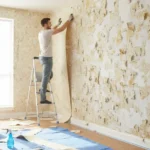 man on a stepladder peeling a large strip of old wallpaper, demonstrating how to remove it easily as part of a DIY guide.