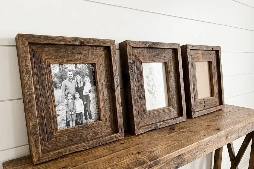 Three rustic, chunky picture frames made from scrap wood standing on a console table, displaying family photos and botanical art against a white shiplap wall.