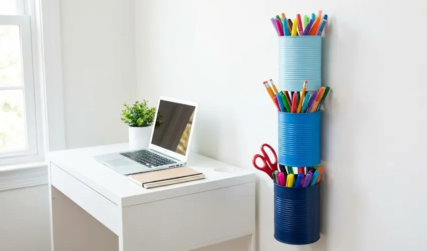 Three painted blue tin cans magnetically attached to a wall in a vertical tower next to a small white desk, filled with markers, pencils, and scissors.