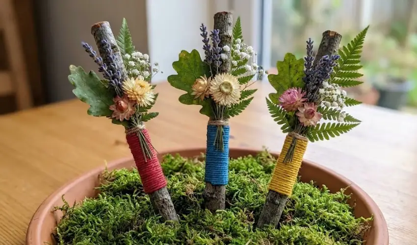 Three handmade mini nature wands crafted from sticks wrapped in colorful yarn and topped with dried flowers and greenery, displayed in a mossy pot.