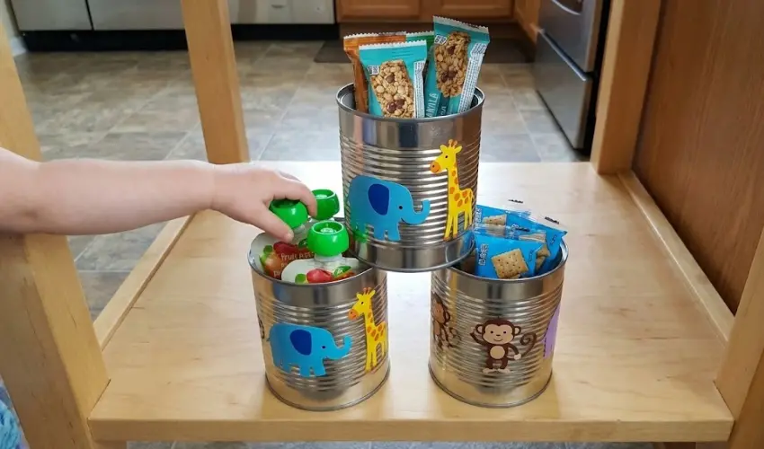 A child reaching for a fruit pouch from a DIY stacked tin can organizer decorated with cute animal stickers and filled with granola bars and crackers.