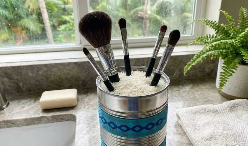 A DIY makeup brush drying rack made from a recycled tin can decorated with blue ribbon and filled with rice, holding wet brushes upright on a bathroom counter.