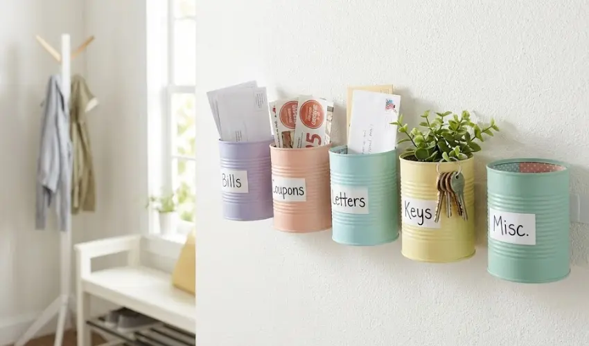 A creative DIY wall-mounted mail sorter made from pastel-painted tin cans labeled for bills, coupons, and letters, displayed in an entryway.