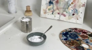 A bowl of baking soda with a spoon placed next to a dirty paint palette and a stained watercolor canvas on a white counter.