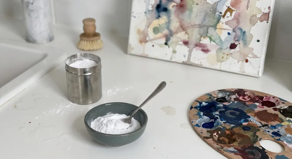 A bowl of baking soda with a spoon placed next to a dirty paint palette and a stained watercolor canvas on a white counter.