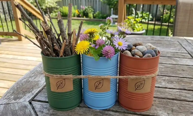 Three painted tin cans in green, blue, and terracotta tied together with twine, labeled with leaf drawings and filled with sticks, wildflowers, and stones on an outdoor wooden table.