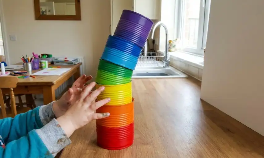 A child’s paint-stained hands balancing a leaning tower of six recycled tin cans painted in rainbow order (red, orange, yellow, green, blue, purple) on a wooden kitchen counter.