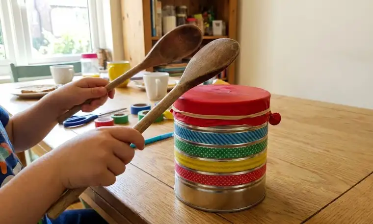 A child playing a DIY tin can drum made with a red balloon stretched over the top and colorful washi tape stripes, using wooden spoons as drumsticks.