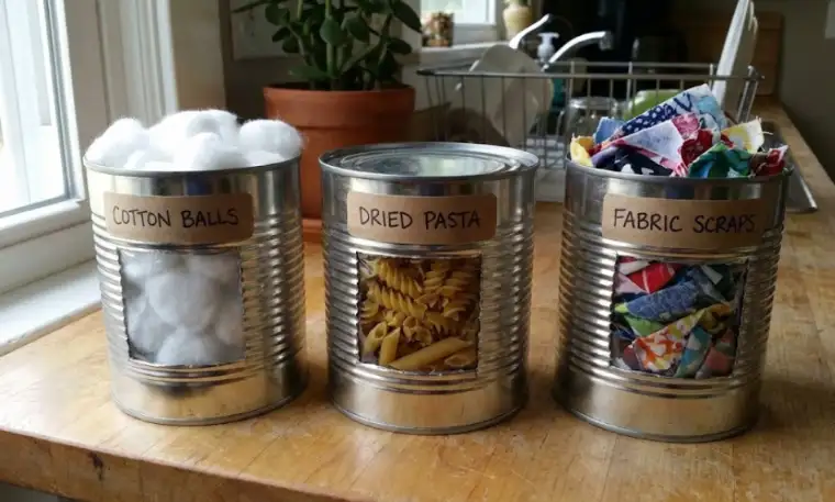Three DIY sensory cans labeled "Cotton Balls," "Dried Pasta," and "Fabric Scraps" sitting on a wooden counter, featuring clear windows that reveal the different textures inside.