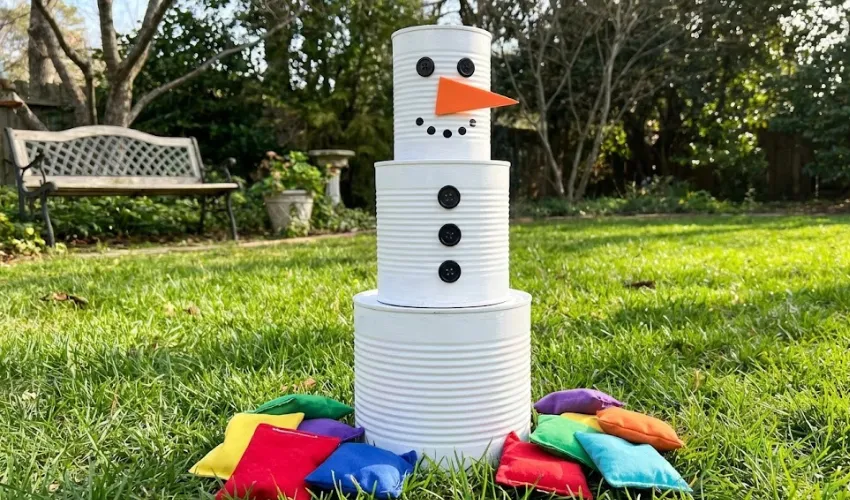 A DIY snowman stacking game made from three white-painted tin cans with black buttons and an orange nose, set up on grass with colorful bean bags.