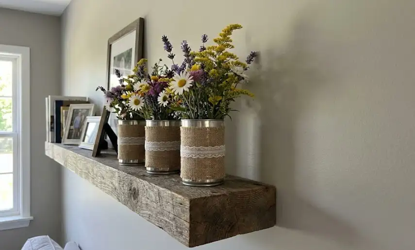 Three upcycled silver tin cans wrapped in brown burlap fabric and white lace trim, arranged in a row on a chunky wooden floating shelf and filled with wild daisies, goldenrod, and purple lavender.