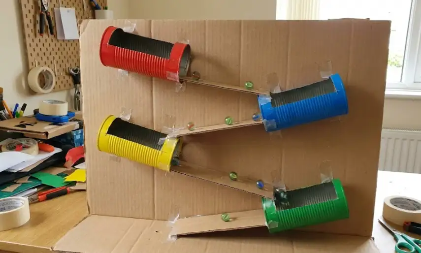 A vertical marble run constructed on a large cardboard sheet, featuring four recycled tin cans (painted red, blue, yellow, green) cut lengthwise to serve as tunnels, connected by zig-zagging cardboard ramps with green glass marbles descending the track.