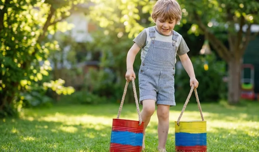 A happy child walking on DIY tin can stilts painted red and yellow in a sunny backyard, demonstrating a fun gross motor activity.