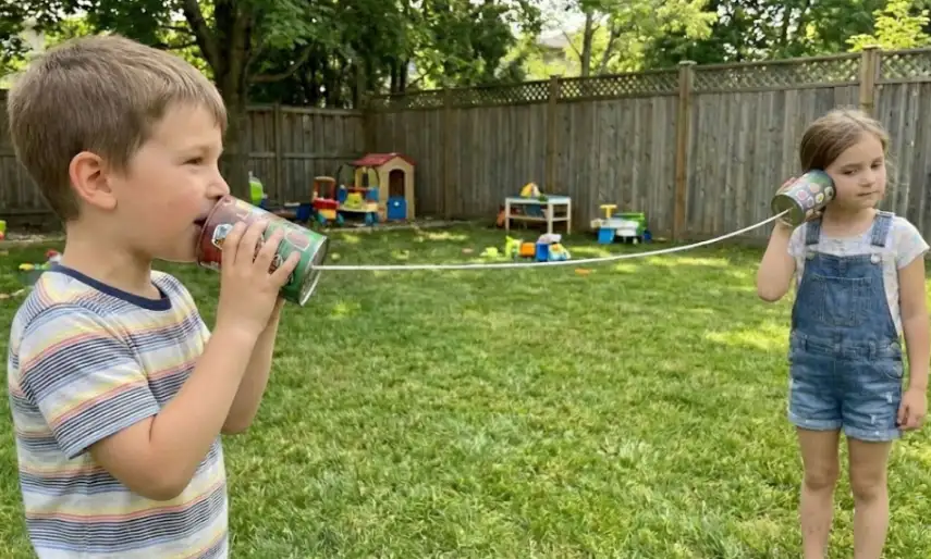 A side-profile shot of a young boy speaking into a green painted tin can while a girl listens through a matching can, connected by a taut white string spanning the distance across a green backyard lawn.