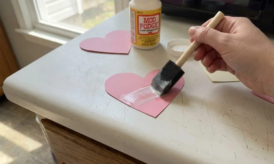 Close-up of a hand using a black foam brush to spread a thin layer of white Mod Podge sealant onto a pink paper heart cutout on a white countertop.