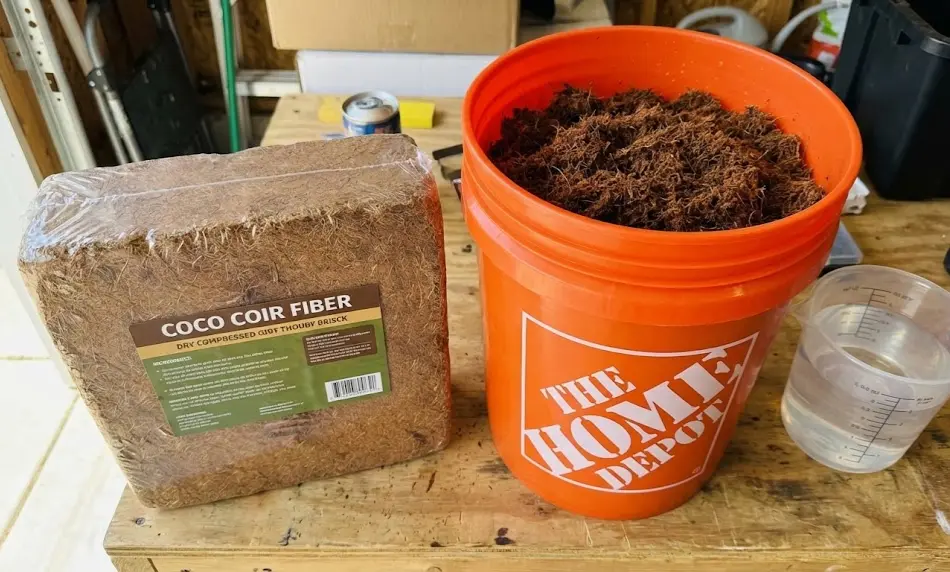 A packaged compressed coco coir fiber brick sitting on a workbench next to an orange 5-gallon bucket filled with expanded, dark brown moist coir and a plastic pitcher of water.