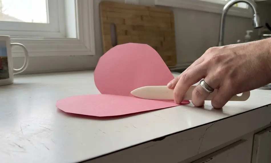 Close-up of a hand using a white bone folder to press a sharp crease into the center of a pink paper heart on a white kitchen countertop.