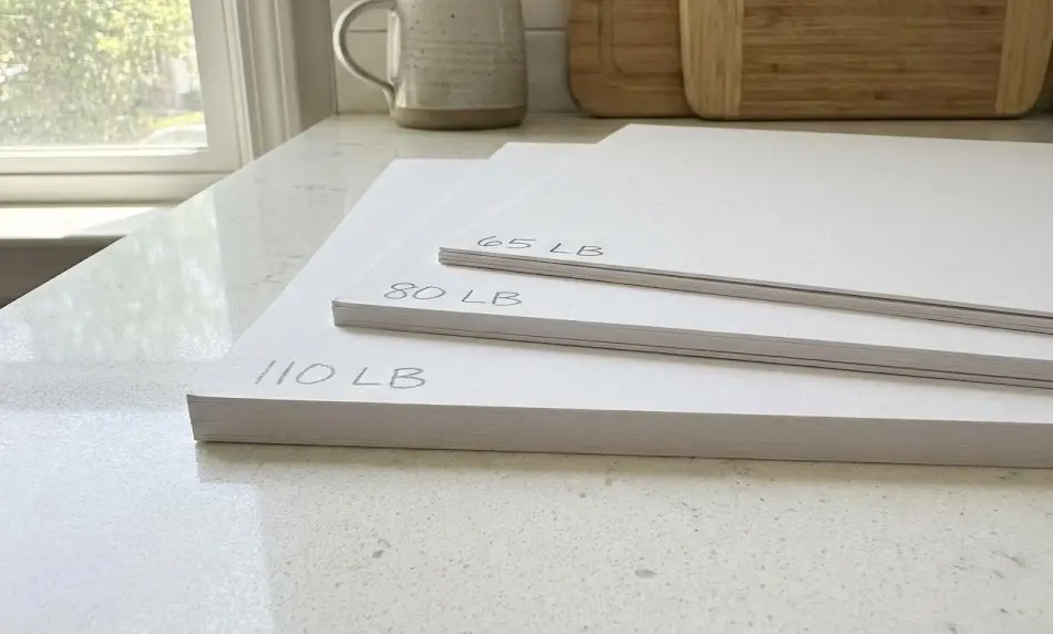 ide-angle view of three white cardstock stacks labeled 65 LB, 80 LB, and 110 LB on a quartz countertop, visually comparing paper thickness for crafting projects.