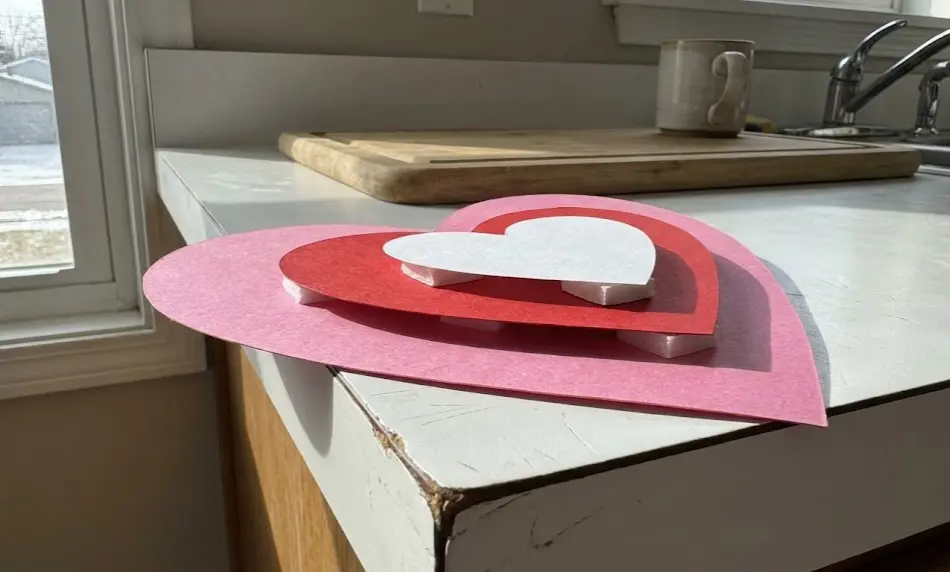 Side-profile view of a 3D Valentine paper craft, displaying pink, red, and white hearts stacked using thick white foam adhesive squares to create elevation and shadow effects.