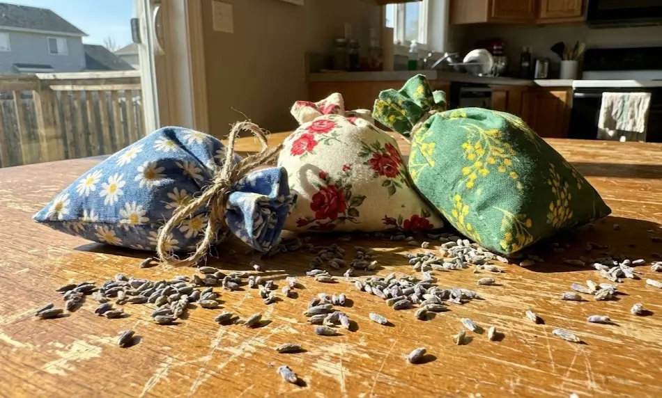 Close-up of three handmade fabric sachets featuring blue, cream, and green floral patterns, tied at the top with rustic twine and resting on a sunlit wooden table surrounded by scattered dried lavender buds.