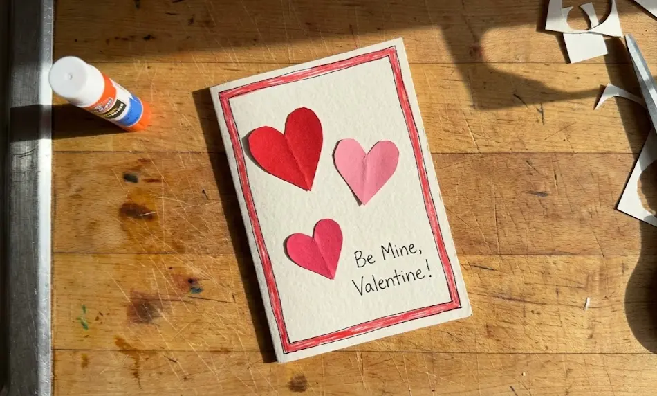A close-up of a handmade Valentine's Day card lying on a wooden workspace, featuring three cut-out paper hearts in red and pink, a hand-drawn red border, and the handwritten phrase "Be Mine, Valentine!" next to a glue stick.