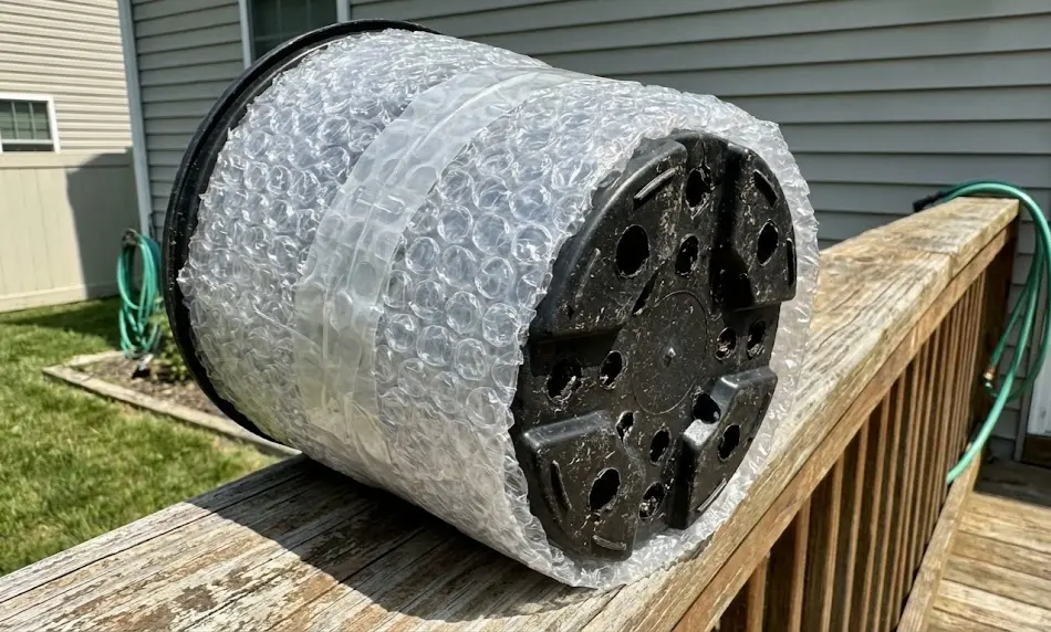 A black plastic nursery pot lying on a wooden deck rail, wrapped in thick layers of clear bubble wrap for winter protection, with the bottom drainage holes clearly visible and uncovered.