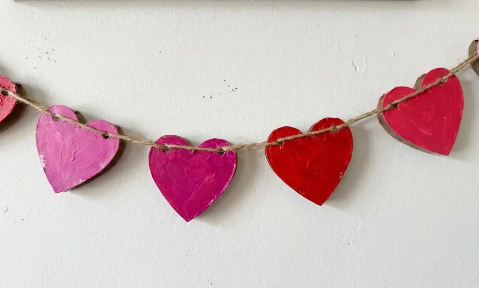 Close-up of a rustic Valentine garland made from painted pink and red cardboard tube hearts threaded onto jute twine against a white wall.