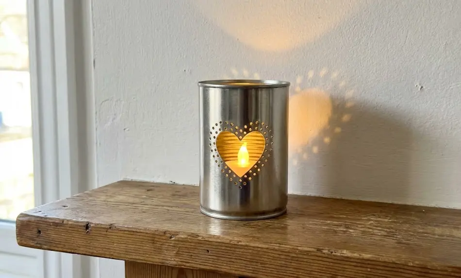 Close-up of a silver recycled tin can with a punched heart design sitting on a rustic wooden shelf, casting a distinct heart-shaped light shadow onto the white wall behind it.