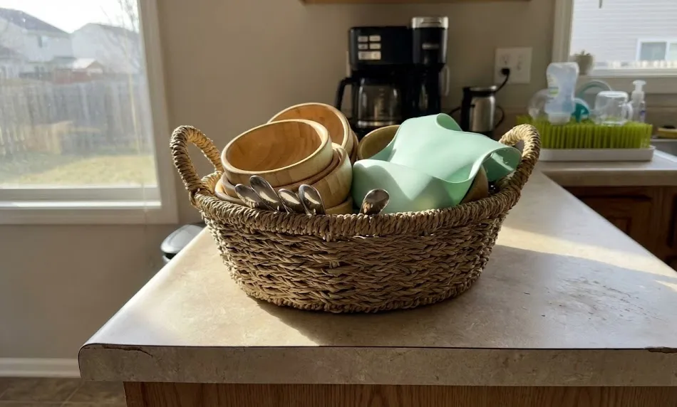 A woven seagrass basket sitting on a kitchen counter, filled with nested bamboo infant bowls, stainless steel spoons, and a mint green silicone bib.